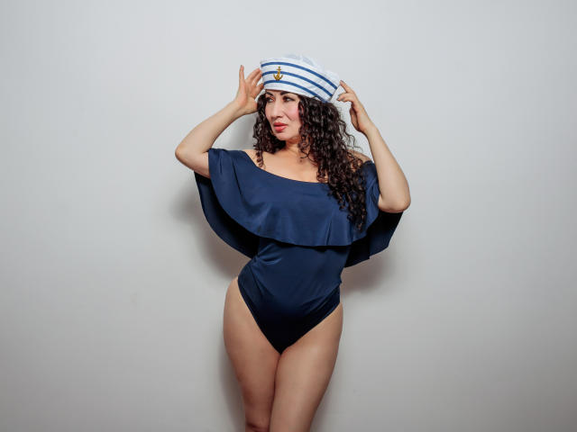 Samantha Antolín, con cabello castaño rizado, vestida con una blusa azul de un solo hombro, pantalones negros de corte alto y un sombrero de rayas estilo marinero, sosteniéndolo en su cabeza. Sus senos son de tamaño medio..