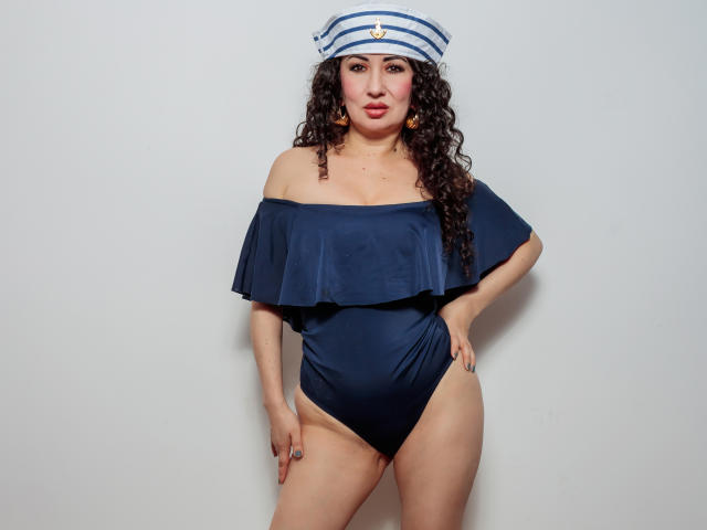 SamanthaAntolin in a navy blue off-shoulder one-piece with ruffle, wearing a sailor hat, curly brown hair, and orange earrings, standing with hands on hips, showing her tits.