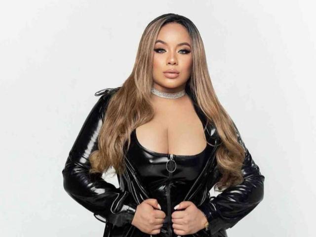 GoddessMiller with long brown hair, wearing a black lace corset, black shorts, and holding a white fur coat. She has large tits, a curvy ass, and a confident look.