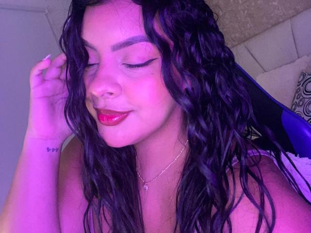 AmarantaCarey with wet black hair, closed eyes, and pink lipstick, covering her tits with one hand, wearing a heart necklace, and nose ring, under purple lighting.