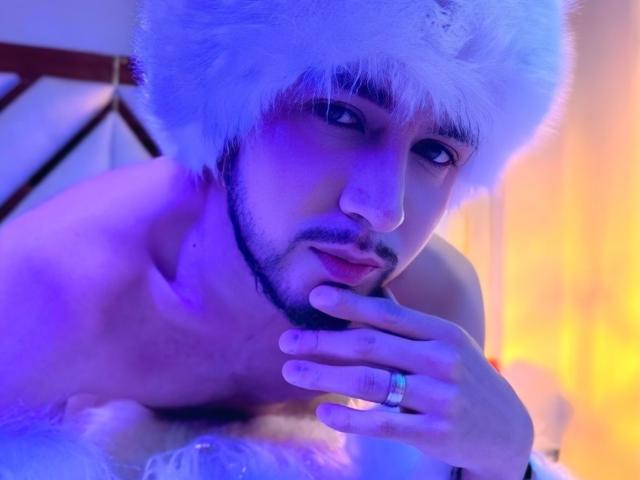Dalton White senza camicia, con un cappello bianco e peloso, illuminazione blu e gialla, che si tocca il mento con un anello al dito, e che guarda seducentemente la telecamera..