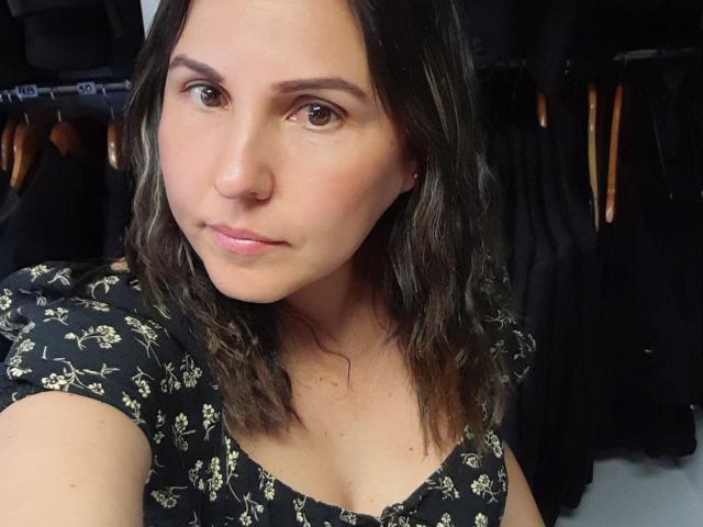 AdelaShery with wavy brown hair, wearing a black floral dress, has a cute nose, subtle pink lips, and brown eyes, standing in a room with clothes in background.