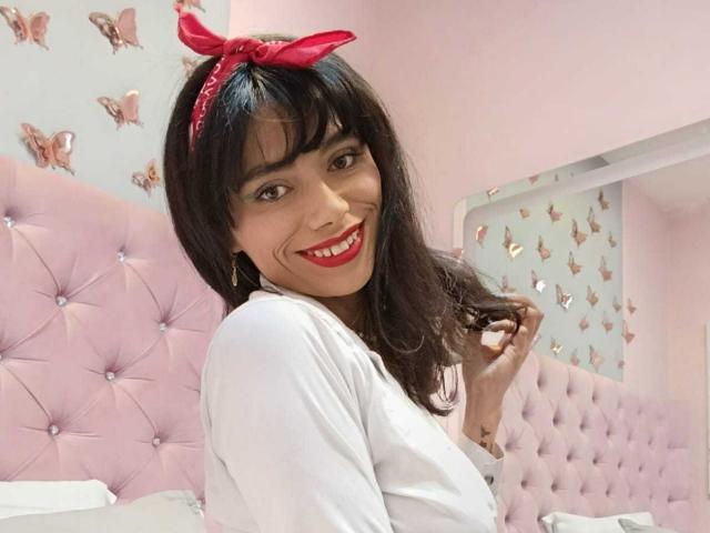 AnnaMooree, con capelli scuri, un fiocco rosso, una camicia bianca, che sorride, si tiene i capelli, un letto rosa con decorazioni, farfalle sul muro, una stanza rosa, rossetto rosso, carina, graziosa, attraente, sexy, felice..
