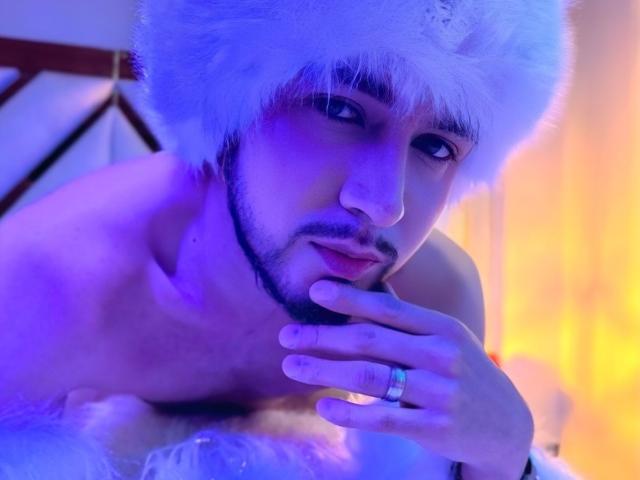 DaltonWhite, torse nu, avec un chapeau blanc et duveteux, éclairage bleu et violet, touchant son menton, montrant une bague à son doigt, souriant légèrement, avec une courte barbe et des cheveux foncés..