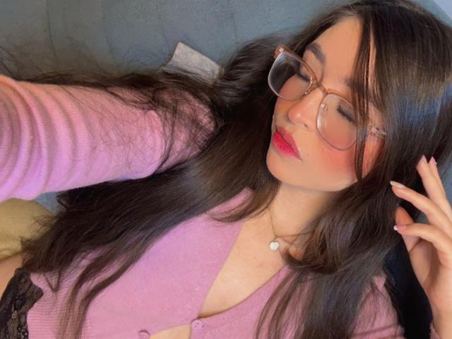EvaMcAllister met lang zwart haar, een roze vestje en een bril, liggend, met gesloten ogen, rode lippenstift, een kleine hartvormige ketting, haar hand op haar hoofd, waarbij haar borsten zichtbaar zijn, ontspannen en mooi..