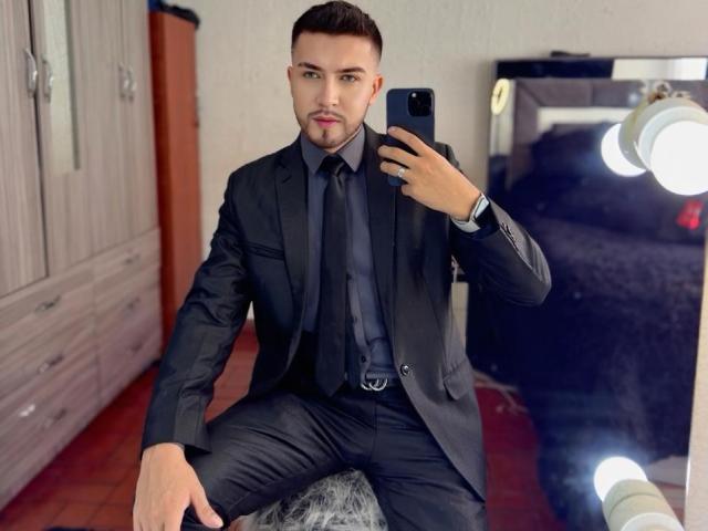 DaltonWhite, gekleed in een zwart pak met een das, maakt een spiegelzelfportret. Hij zit op een pluizige grijze kruk, houdt een telefoon vast en kijkt zelfverzekerd..