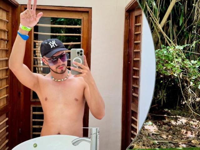 Naked DaltonWhite met een baseballpet, zonnebril en een ketting, die een vrede-teken maakt, een telefoon vasthoudt, een blauwe polsband draagt, in een selfie voor een spiegel in een badkamer..