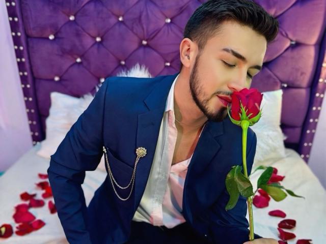 DaltonWhite, con un abito blu scuro e una camicia rosa, si sporge in avanti, baciando una rosa rossa, i petali sparsi su un letto, con una testiera viola imbottita sullo sfondo..