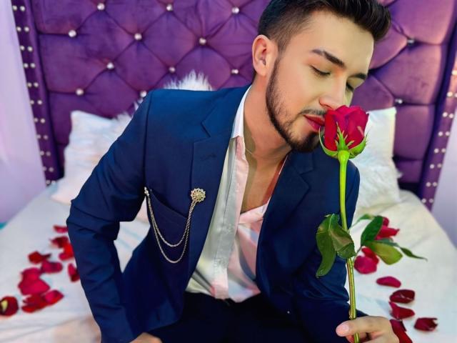 DaltonWhite con una rosa roja en la boca, vistiendo un traje azul, camisa blanca y una cadena, sentado en una cama con pétalos de rosa, con un cabecero morado acolchado en el fondo..