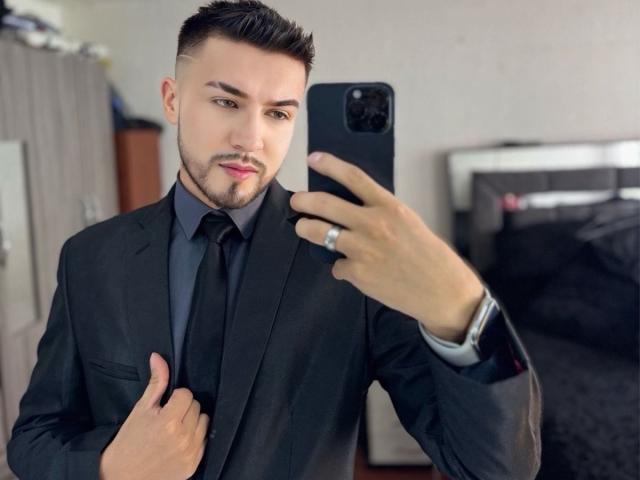DaltonWhite, vêtu d'un costume noir avec une cravate noire, tenant un smartphone et se prenant un selfie devant un miroir. Il a les cheveux courts et foncés, une barbe soignée, et porte une bague à la main droite..