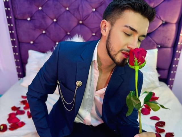 DaltonWhite draagt een marineblauw pak met een roze shirt, en houdt een rode roos voor zijn neus. Hij zit op een bed met rozenblaadjes. Hij heeft een baard en kort donker haar..