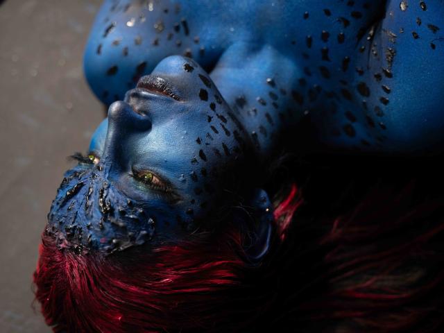 SofyaLane, avec du maquillage bleu sur le visage, des taches noires et des cheveux roux, allongée, regardant vers le haut, avec des yeux verts, une peau humide et une bouche légèrement ouverte. Seins visibles..