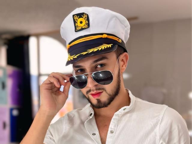 DaltonWhite usando um chapéu branco de capitão com um emblema amarelo, ajustando óculos de sol espelhados e sorrindo com lábios vermelhos, vestindo uma camisa branca de botões..