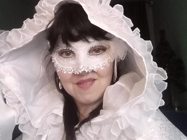 IllusionGold with white lace mask and ruffled hood, smiling. She's got dark brown hair, pink lipstick, and diamond earrings. Her tits are partly visible under the white fabric.
