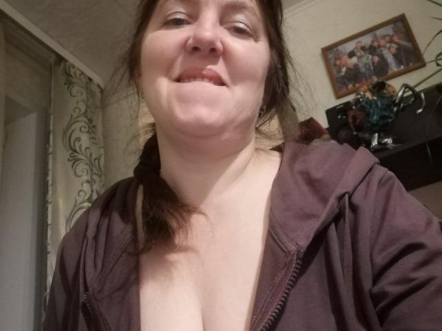 IllusionGold with pale skin, brown hair in a loose ponytail, smiling slightly, wearing a brown zip-up, showing cleavage, in a casual indoor setting.