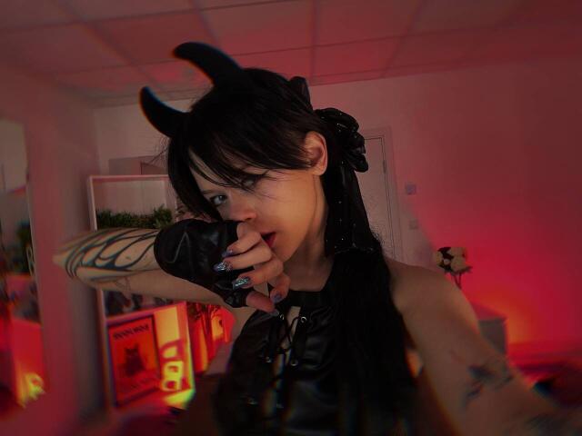 SimaWendy with black devil horns, black leather top, fingerless gloves, tattoos, and long black hair, biting her finger, red and orange lighting, focused selfie.