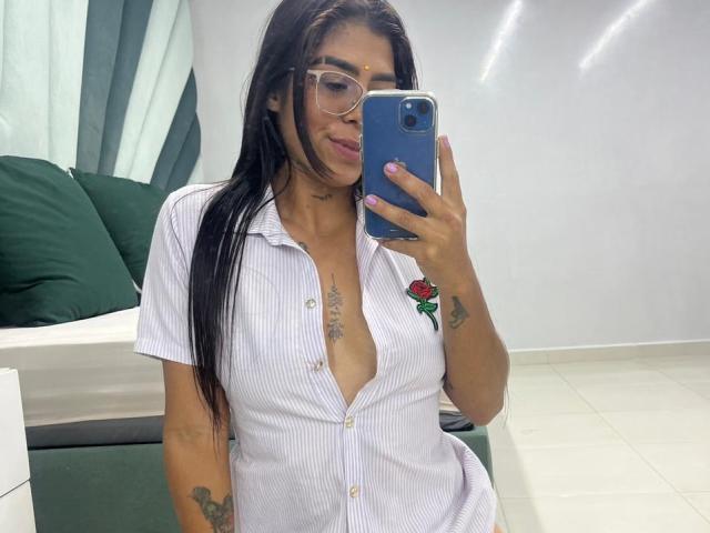 VioletaGates with long black hair, glasses, and a low-cut, button-down, white and pink striped shirt, showing her tits. She's holding a blue phone, smiling, with tattoos visible.