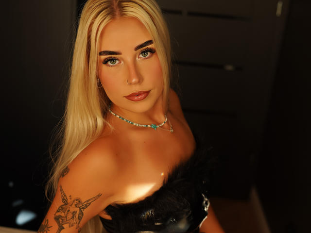 Blonde KristyQueen with feathery black top, blue necklace, tattoo on right arm, looking up, green eyes, dark lipstick, and slight smile, standing in dimly lit room.