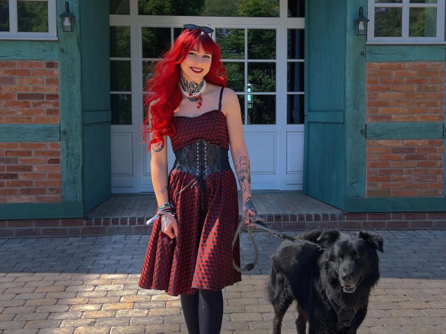 Teufelshelferin-hot with bright red hair, wearing a checkered dress, black corset, and tattoos, holds a black dog's leash. She's smiling, standing on a brick pathway.