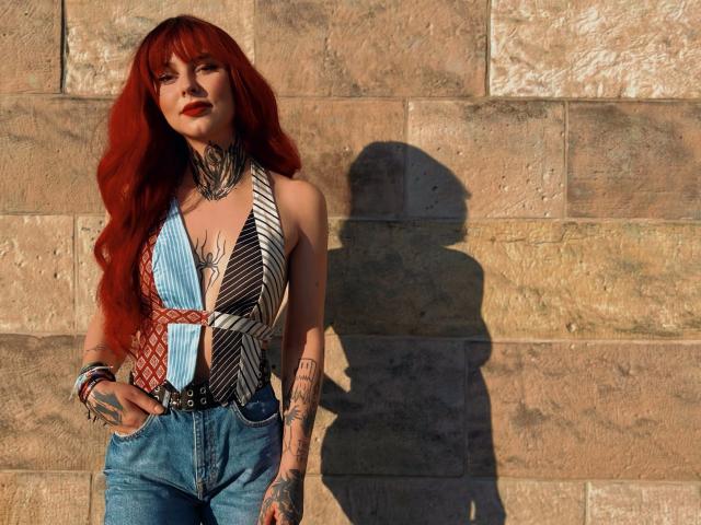 Teufelshelferin-hot has red hair, a deep V-neck patterned top, tattoos, and wears blue jeans. Her shadow is on the wall, hands in pockets, looking confident.