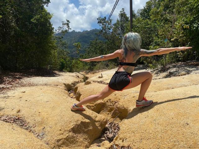 Prof-Paula-Bunt-hot with blue hair, black sports bra, and black shorts, is squatting on yellow rocks, stretching arms wide, showing her ass and tattooed back in a sunny forest.