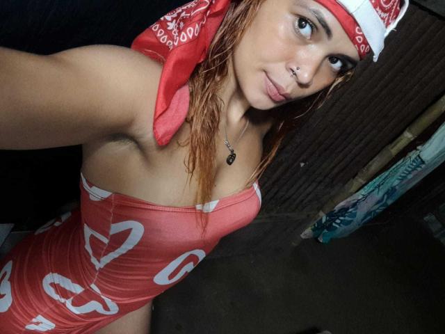 DomiEvans con un pañuelo rojo, cabello mojado, piercing en la nariz y un vestido rojo con un corazón blanco, mostrando sus senos, un pequeño collar con forma de corazón y una expresión de labios fruncidos en una habitación con poca luz..