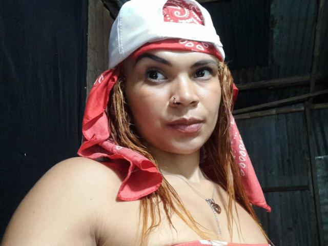 DomiEvans, com um lenço vermelho na cabeça, boné branco, piercing no nariz e colar, mostrando pele bronzeada e olhos castanhos, olhando de lado com um leve sorriso, e ombros descobertos..