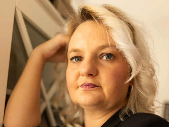 Hotblondi-hot with blonde, wavy hair and white streak, blue eyes, light skin, wearing black shirt, hand in hair, slight smile, soft pink lipstick, looking forward.