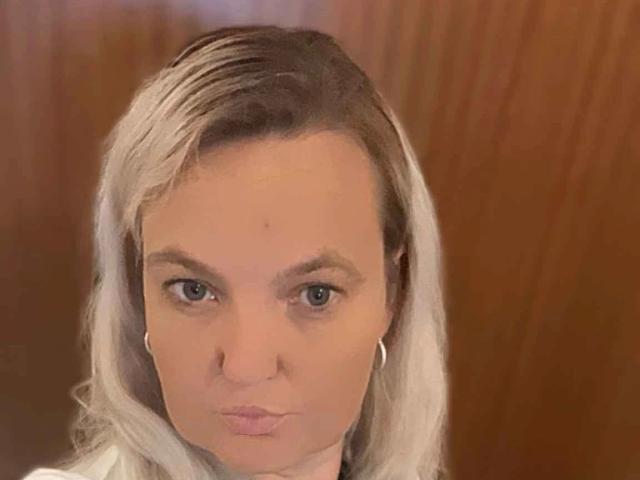 Hotblondi-hot has blonde hair, blue eyes, and a small mole on her face, with pouty pink lips and white ear hoops, looking straight at the camera.