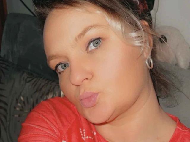 Hotblondi-hot with pouty lips, blue eyes, and silver hair in a messy updo, wearing a red shirt, has small tits, and is making a silly face.