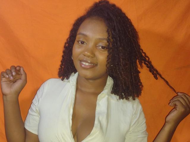 BarbaraSweetz with dark curly hair, smiling, wearing a white shirt, holding her hair with one hand, showing her chest, orange background, medium skin, cute, attractive.
