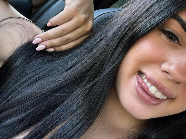 KimberlyGarx's face with long black hair, smiling with pink lips, white teeth, and a hand touching her hair. She has light brown skin and pink nail polish.