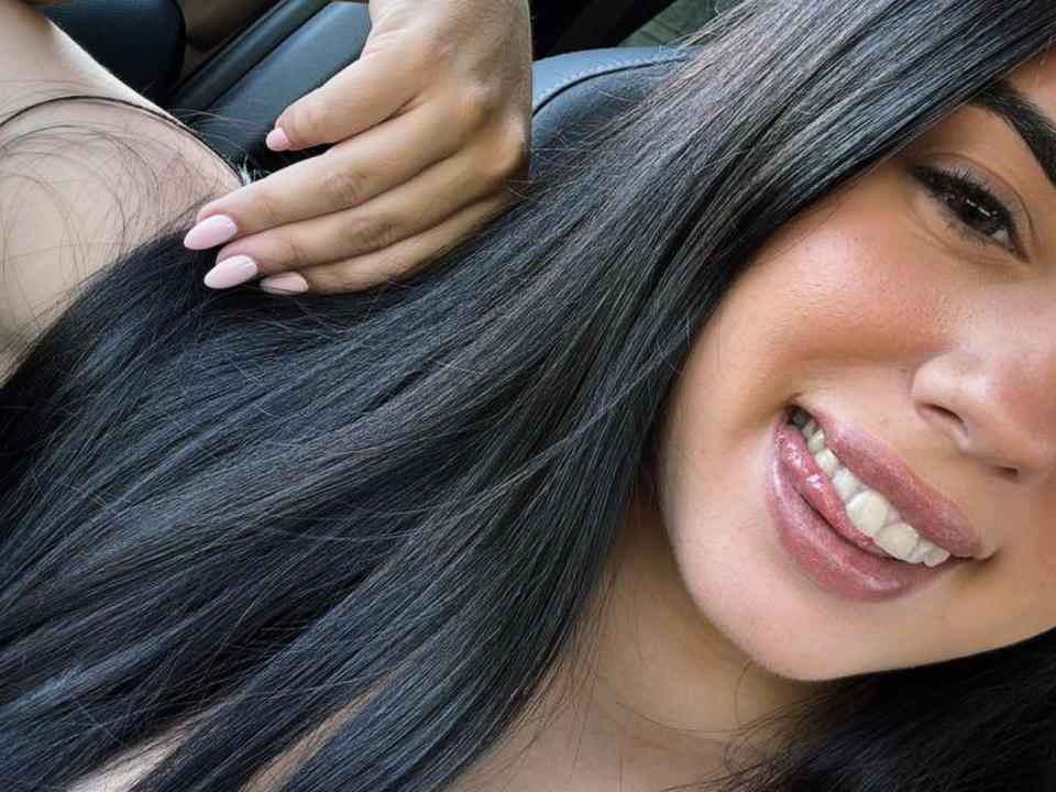 KimberlyGarx's face with long black hair, smiling with pink lips, white teeth, and a hand touching her hair. She has light brown skin and pink nail polish.