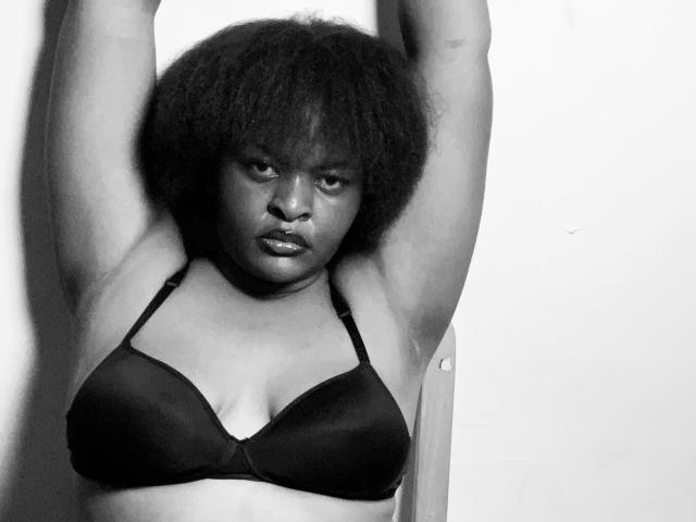 Black and white photo of SpiceBomb with dark, curly hair, arms raised, wearing a black bra, showing small to medium-sized tits, serious expression, standing against a plain wall.
