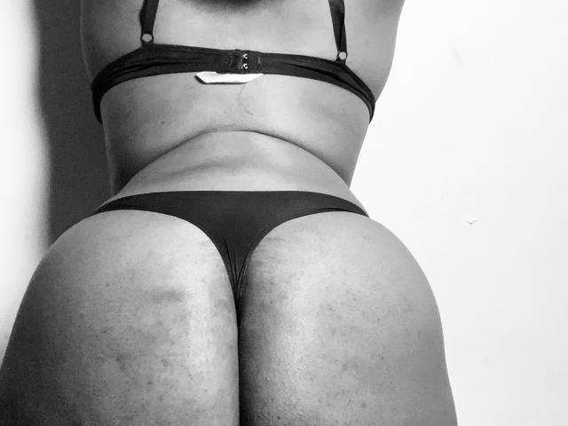 SpiceBomb's black bra and thong, showing her back and butt. She's leaning forward, highlighting her rounded ass and slightly textured skin. Black and white photo.