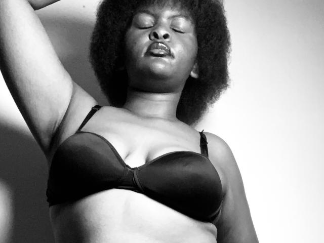 SpiceBomb with closed eyes, dark curly afro, wearing a black bra, arm raised, showing medium-sized tits, soft lighting, black and white, slightly open mouth, relaxed expression.