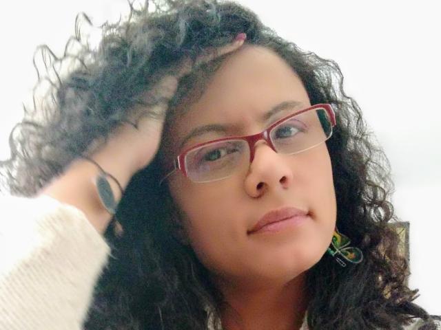 RosannaBlue with curly black hair, red glasses, and a nose ring, has a hand on her head, looking slightly annoyed, wearing a white sweater, showing a tattoo on her wrist.