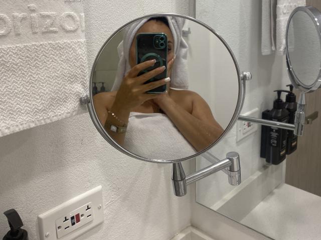 GalaMalkova with a towel on her head, holding a phone in front of a round mirror, showing her bare tits. Bathroom, mirror, towel, black bottles in background.