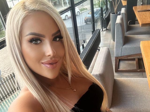 MissLeylaBlond with long blonde hair, green eyes, full pink lips, and big tits, wearing a black strapless top, smiling in a cafe.
