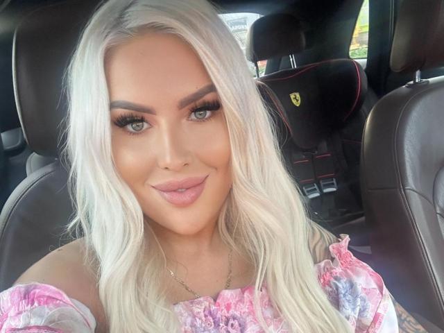 MissLeylaBlond with platinum blonde hair, full lips, and thick eyebrows, wearing a pink floral dress, sitting in a car with a Ferrari seat. Her tits are small.