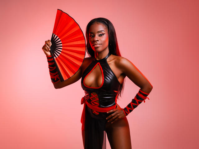 MiaHopson with dark skin, long black hair, holding a red fan, wearing a black leather dress with red trim, showing her tits, and red ribbon wristbands.
