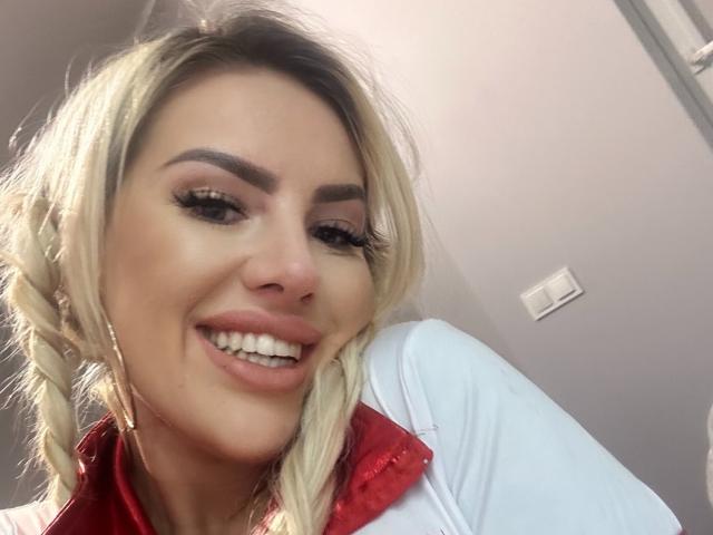MissLeylaBlond has blonde braids, thick eyebrows, long eyelashes, and is smiling with her pink lips showing. She's wearing a red and white shirt.