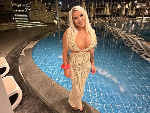 MissLeylaBlond with long blonde hair, large boobs, in a cream crochet dress, standing by a blue-tiled pool, wearing a pink scrunchie and black flip-flops, smiling.