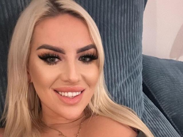 MissLeylaBlond has blonde hair, thick eyebrows, long eyelashes, and is smiling with her white teeth. She's wearing a gold necklace, sitting on a blue ribbed couch.