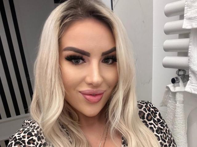 MissLeylaBlond has blonde hair, thick eyebrows, long lashes, and pink lips. She's wearing a leopard print top, and a white towel is on the right.