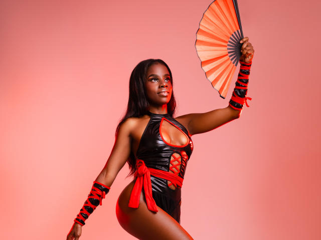 MiaHopson with dark skin, long black hair, wearing a black leather halter dress with red lace, holding an orange fan, and red wristbands, posing confidently.