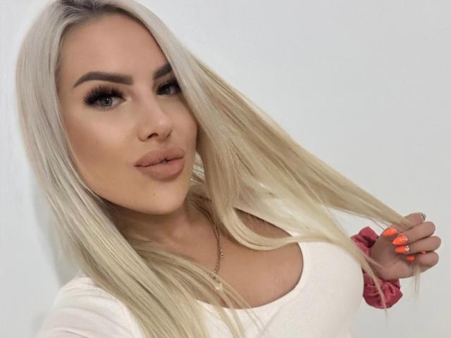 MissLeylaBlond has blonde hair, full tits, and is pouting with pink lips. She's wearing a white top and has orange nails, holding her hair with a pink scrunchie.