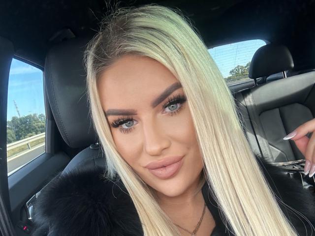 Blond MissLeylaBlond with full lips, long eyelashes, and fur collar in a car, smiling at the camera, showing her blue eyes and dark eyebrows.