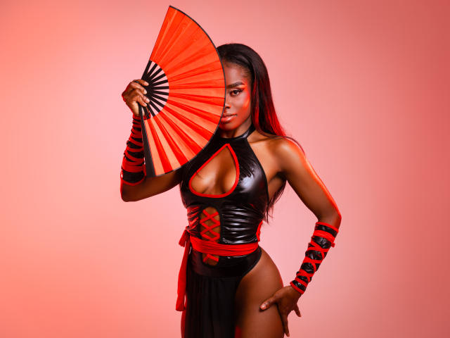 MiaHopson with dark skin, wearing a black leather dress, red and black wristbands, holding a red and black fan, covering her tits, pink background, long red hair.