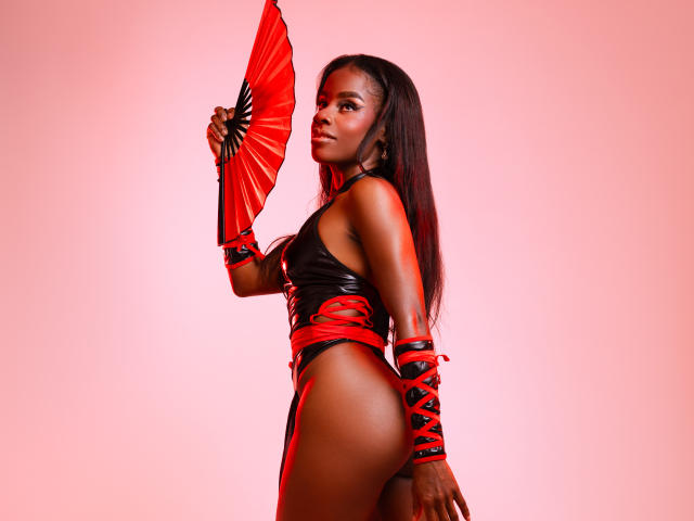MiaHopson with dark skin, long black hair, wearing a red and black latex outfit, holding a red fan, showing her ass, looking over her shoulder with a confident expression.